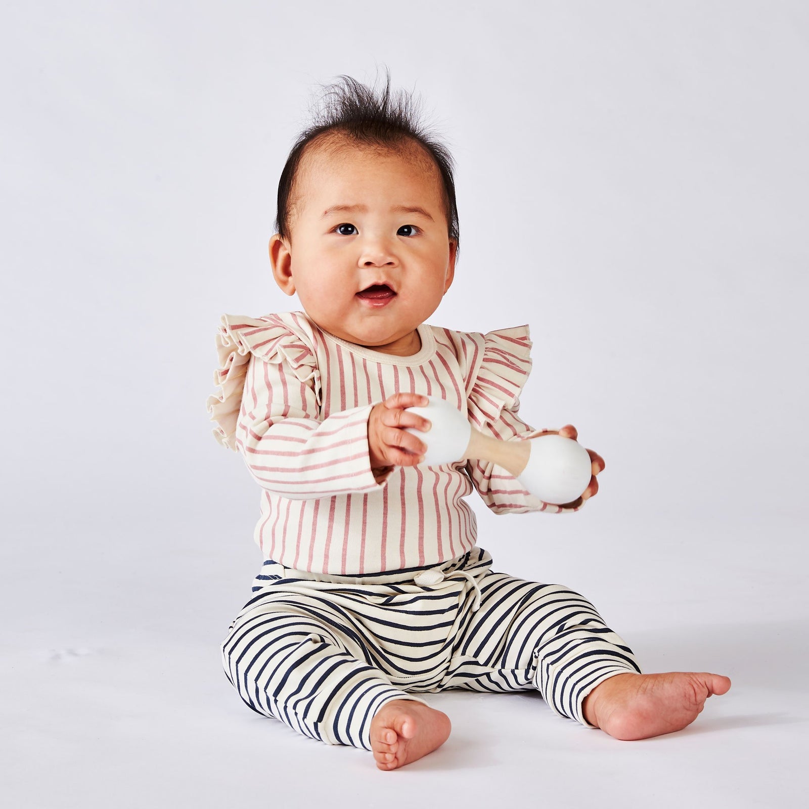 Baby wearing Pehr Stripes Away Dark Pink with Ruffle Organic One-Piece, Long Sleeve and Pehr Stripes Away Bloomers Ink Blue Organic Bloomers & Shorts. GOTS Certified Organic Cotton & Dyes. White with dark pink stripes, long sleeve, ruffles on shoulders, button closure at bottom.