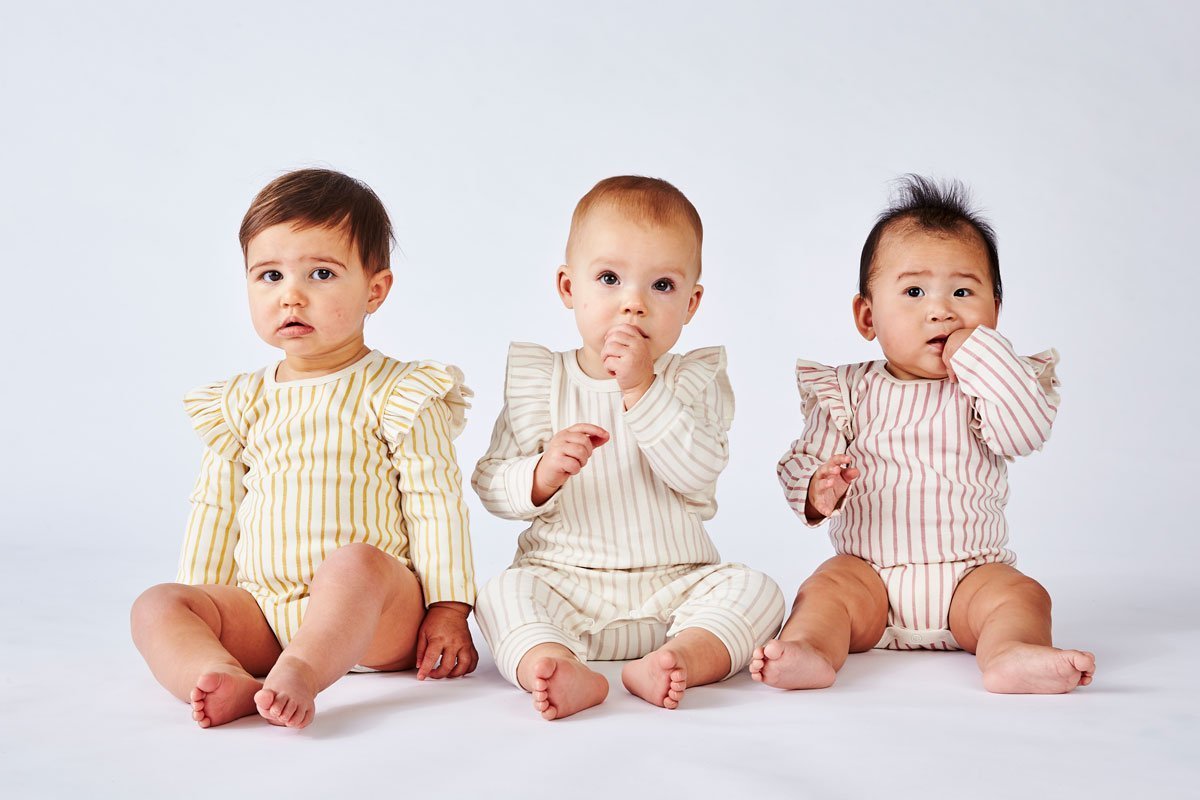 Three babies wearing Pehr Stripes Away Marigold, Pebble Grey, and Dark Pink with Ruffle Organic One-Piece, Long Sleeve. GOTS Certified Organic Cotton & Dyes. White with stripes, long sleeve, ruffle on shoulders, button closure at bottom.